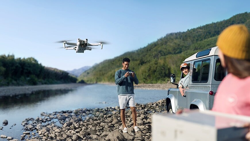 Dron DJI Mini 3 Fly More Combo CP.MA.00000613.01 widok na mężczyznę siedzącego w samochodzie oraz na mężczyznę stojącego na kamieniach w trakcie sterowania latającym nad nim dronem